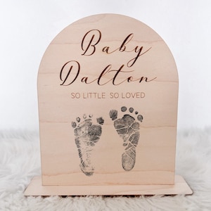 May include: A wooden arch-shaped sign with the text "Baby Dalton" and "So Little So Loved" in a cursive font. Two black and white baby footprints are printed on the sign.