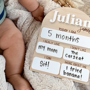 以下が含まれることがあります： 「Julian james」と「TODAY I AM 5 months」という白い文字が書かれた木製のマイルストーンボード。ボードには、白いテキストボックスがあり、次のようなテキストが書かれています。「I REALLY LOVE my mom」、「I CAN Sit!」、「I DON'T LIKE The Carseat」、「MORE ABOUT ME I tried banana!」