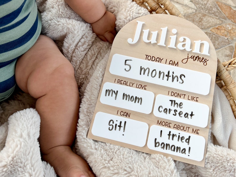 以下が含まれることがあります： 「Julian james」と「TODAY I AM 5 months」という白い文字が書かれた木製のマイルストーンボード。ボードには、白いテキストボックスがあり、次のようなテキストが書かれています。「I REALLY LOVE my mom」、「I CAN Sit!」、「I DON'T LIKE The Carseat」、「MORE ABOUT ME I tried banana!」
