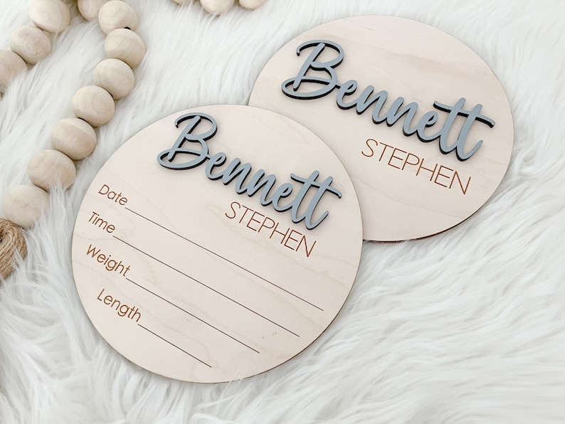 K&ouml;nnte beinhalten: Zwei runde Holzschilder zur Babyank&uuml;ndigung. Jedes Schild tr&auml;gt den Namen "Bennett" in grauem 3D-Schriftzug, darunter "Stephen" in kleinerer Schrift. Ein Schild hat Linien f&uuml;r Datum, Uhrzeit, Gewicht und L&auml;nge.