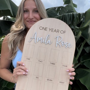 A wooden birthday photo board with the text "ONE YEAR OF Amila Rose" in white script. Showing the small size photo board. The board has twelve rectangular spaces labeled with numbers one through twelve. Photos needed for this size photo board are wallet size.