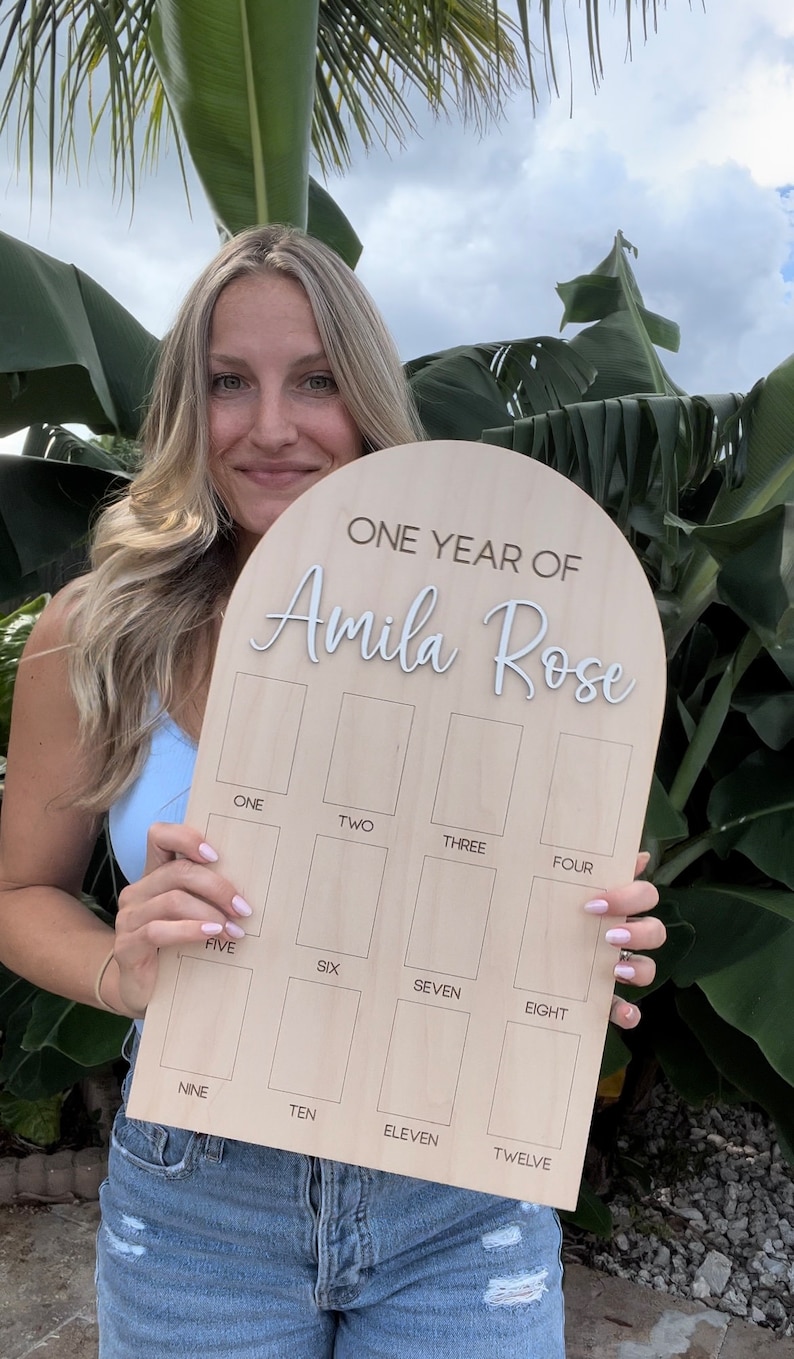 A wooden birthday photo board with the text "ONE YEAR OF Amila Rose" in white script. Showing the small size photo board. The board has twelve rectangular spaces labeled with numbers one through twelve. Photos needed for this size photo board are wallet size.
