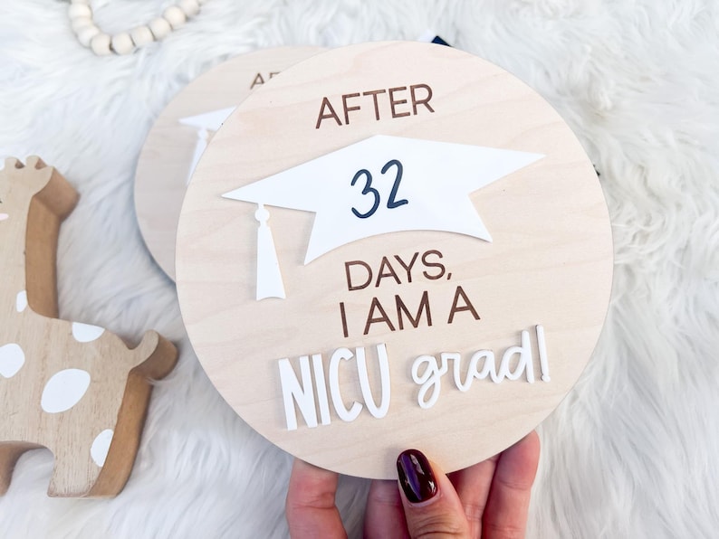 May include: A wooden round sign with the text "After 32 days, I am a NICU grad!" in white and brown lettering. A white graduation cap is on top of the number 32.