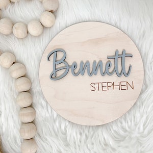 K&ouml;nnte beinhalten: Runder Holzschild mit dem Namen "Bennett" in grauer Schrift, &uuml;ber dem Holz angeordnet. Der Name "Stephen" ist in kleinerer, rotbrauner Schrift darunter gedruckt. Das Schild ist aus hellem Holz.