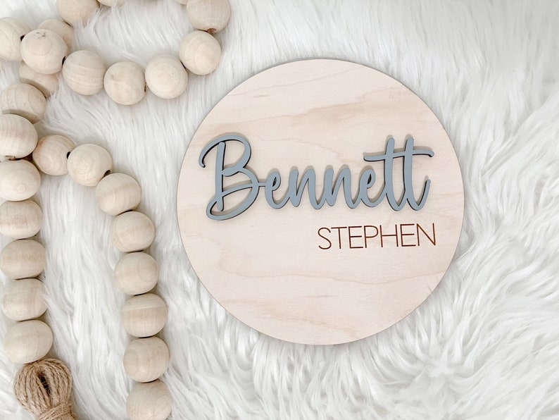 K&ouml;nnte beinhalten: Runder Holzschild mit dem Namen "Bennett" in grauer Schrift, &uuml;ber dem Holz angeordnet. Der Name "Stephen" ist in kleinerer, rotbrauner Schrift darunter gedruckt. Das Schild ist aus hellem Holz.