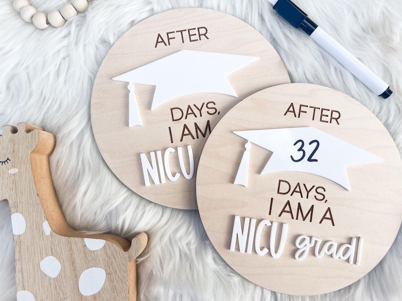 May include: Two wooden round signs with a white graduation cap on top. The signs are decorated with the text "After" and "Days, I am a NICU grad". The sign on the left says "After Days, I am NICU".