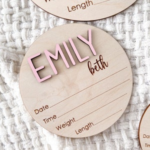 Wooden round baby announcement plaque with the name "EMILY" in pink 3D letters and the name "beth" in script. The plaque includes lines for date, time, weight, and length. Other similar plaques are visible.