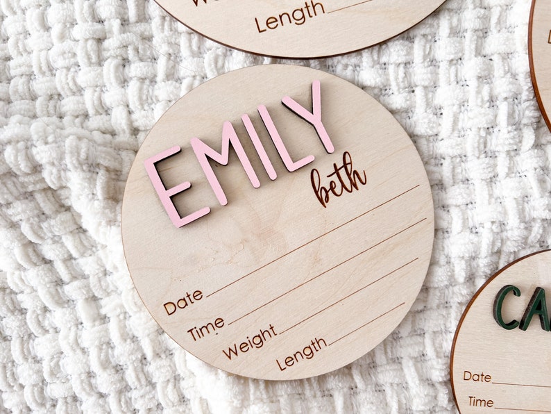 Wooden round baby announcement plaque with the name "EMILY" in pink 3D letters and the name "beth" in script. The plaque includes lines for date, time, weight, and length. Other similar plaques are visible.