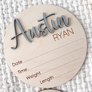 K&ouml;nnte beinhalten: Ein runder Geburtsanzeiger aus Holz mit dem Namen "Austin" in blauer Schrift und dem Namen "Ryan" in braunen Blockbuchstaben. Das Schild hat Linien f&uuml;r Datum, Uhrzeit, Gewicht und L&auml;nge. Der Hintergrund ist eine wei&szlig;e, strukturierte Oberfl&auml;che.