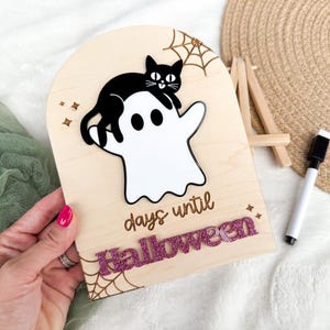 K&ouml;nnte beinhalten: H&ouml;lzernes Halloween-Countdown-Schild mit einer schwarzen Katze auf einem wei&szlig;en Geist. Das Schild zeigt "Tage bis Halloween" mit glitzernden rosa Buchstaben. Enth&auml;lt ein Spinnennetz-Design und Sternakzente.