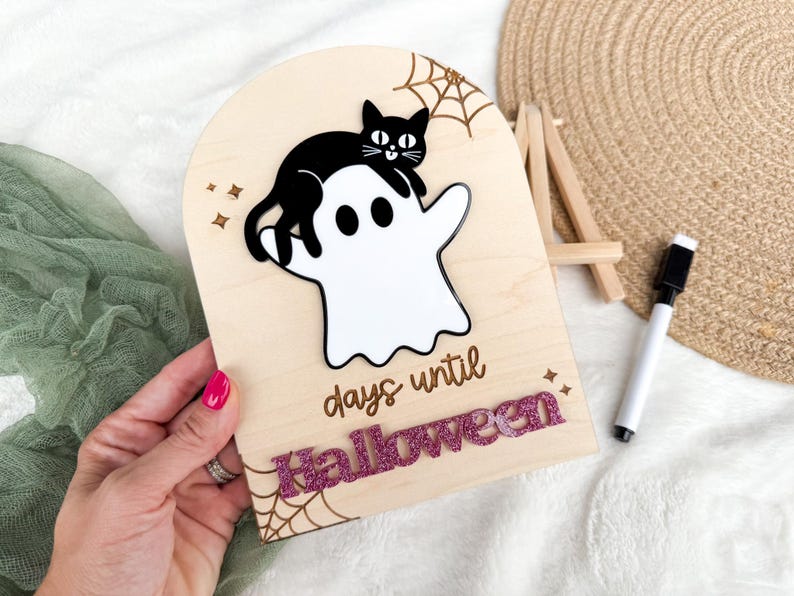 K&ouml;nnte beinhalten: H&ouml;lzernes Halloween-Countdown-Schild mit einer schwarzen Katze auf einem wei&szlig;en Geist. Das Schild zeigt "Tage bis Halloween" mit glitzernden rosa Buchstaben. Enth&auml;lt ein Spinnennetz-Design und Sternakzente.