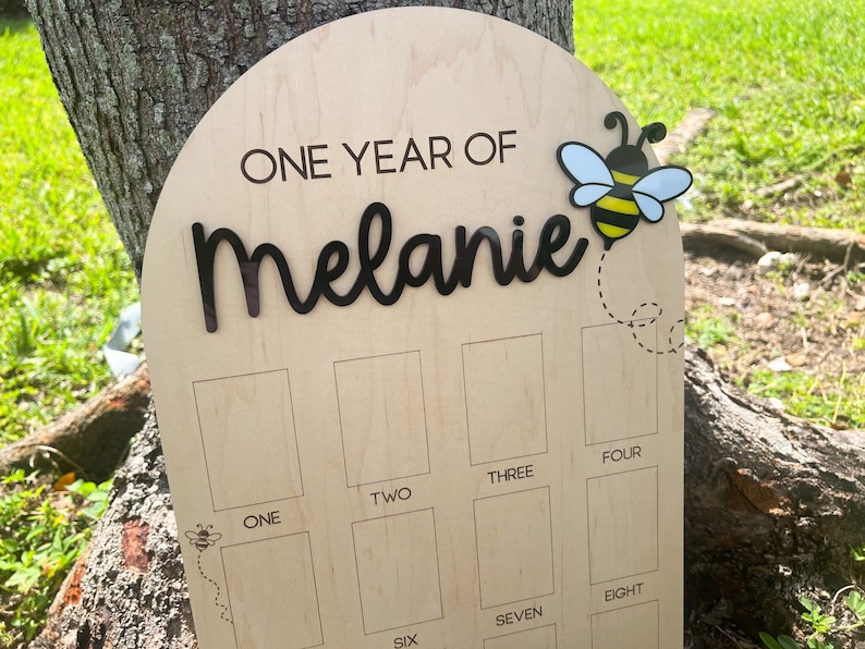Op de afbeelding: Een boogvormig mijlpaalbord van lichtgekleurd hout met de tekst "ONE YEAR OF Melanie" in zwarte letters. Een 3D-bijenicoon is rechtsboven bevestigd met een gestippelde vliegroute. Daaronder zijn twaalf rechthoekige fotovakken gegraveerd.