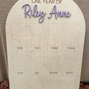 A wooden milestone board with an arched top. The board features the text "ONE YEAR OF Riley Anne" in a purple font. Below are twelve rectangular spaces labeled with the numbers one through twelve. The text "NAME IN VIOLET" is in the upper right corner.
