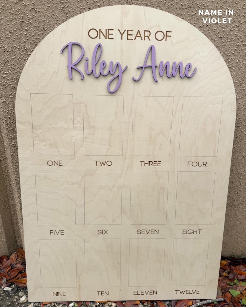 A wooden milestone board with an arched top. The board features the text "ONE YEAR OF Riley Anne" in a purple font. Below are twelve rectangular spaces labeled with the numbers one through twelve. The text "NAME IN VIOLET" is in the upper right corner.