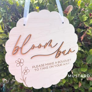 Pu&ograve; includere: Un cartello in legno bianco con bordi a festoni e il testo "Bloom Bar" in corsivo. Il cartello include anche il testo "Please make a bouquet to take on your way" e un disegno floreale.