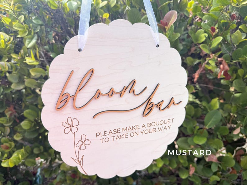 Pu&ograve; includere: Un cartello in legno bianco con bordi a festoni e il testo "Bloom Bar" in corsivo. Il cartello include anche il testo "Please make a bouquet to take on your way" e un disegno floreale.