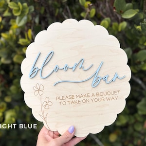 Pu&ograve; includere: Un cartello in legno blu chiaro con il testo "bloom bar" in un font corsivo. Il cartello ha anche il testo "Please make a bouquet to take on your way" in un font pi&ugrave; piccolo.