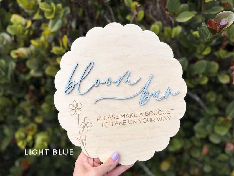 Pu&ograve; includere: Un cartello in legno blu chiaro con il testo "bloom bar" in un font corsivo. Il cartello ha anche il testo "Please make a bouquet to take on your way" in un font pi&ugrave; piccolo.