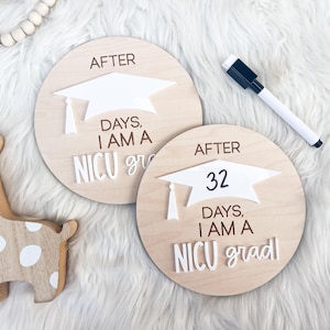May include: Two wooden signs with white graduation caps and the text "After [number] days, I am a NICU grad". The signs are on a white fuzzy surface.