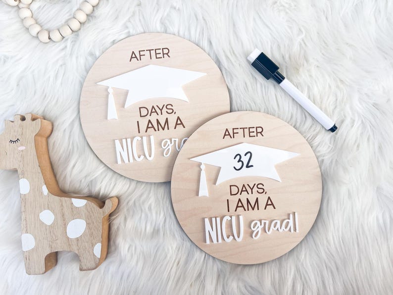 May include: Two wooden signs with white graduation caps and the text "After [number] days, I am a NICU grad". The signs are on a white fuzzy surface.