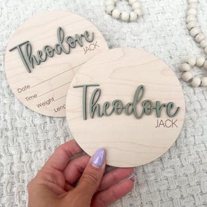 K&ouml;nnte beinhalten: Zwei runde Holzschilder zur Babyank&uuml;ndigung. Der Name "Theodore Jack" ist in einer kursiven Schrift in einem ged&auml;mpften Gr&uuml;nton geschrieben. Ein Schild hat Linien f&uuml;r Datum, Uhrzeit, Gewicht und L&auml;nge. Die Schilder liegen auf einer wei&szlig;en, strukturierten Oberfl&auml;che.