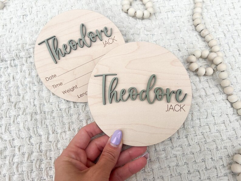K&ouml;nnte beinhalten: Zwei runde Holzschilder zur Babyank&uuml;ndigung. Der Name "Theodore Jack" ist in einer kursiven Schrift in einem ged&auml;mpften Gr&uuml;nton geschrieben. Ein Schild hat Linien f&uuml;r Datum, Uhrzeit, Gewicht und L&auml;nge. Die Schilder liegen auf einer wei&szlig;en, strukturierten Oberfl&auml;che.