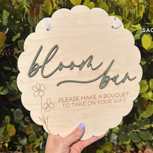 Pu&ograve; includere: Un cartello di legno con un bordo a festoni, dipinto in un colore legno chiaro. Il cartello dice "Bloom Bar" in scrittura verde e "Per favore, fai un bouquet da portare via" in testo marrone.