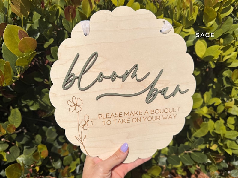 Pu&ograve; includere: Un cartello di legno con un bordo a festoni, dipinto in un colore legno chiaro. Il cartello dice "Bloom Bar" in scrittura verde e "Per favore, fai un bouquet da portare via" in testo marrone.