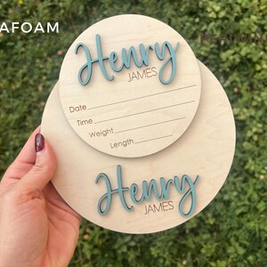 May include: Two round wooden baby announcement signs. The top sign has lines for date, time, weight, and length. The name "Henry James" is written in teal script on both signs. The word "SEAFOAM" is in the top left corner.
