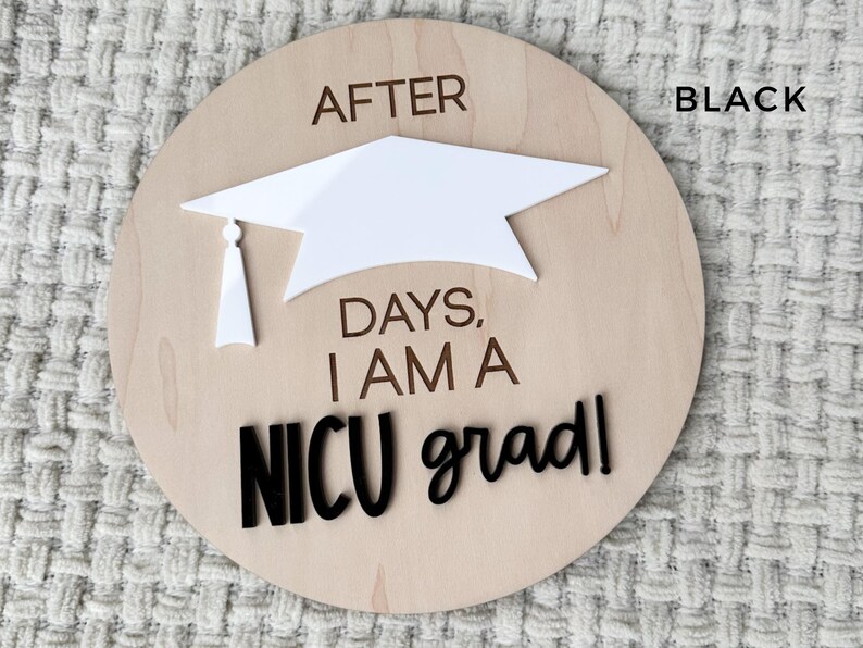May include: A wooden round sign with the text "After ____ days, I am a NICU grad!" in black lettering. A white graduation cap is attached to the sign.