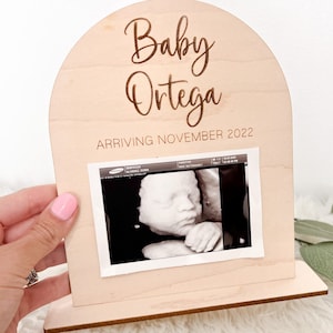 May include: A wooden arch-shaped sign with the text "Baby Ortega Arriving November 2022" and a black and white ultrasound image.