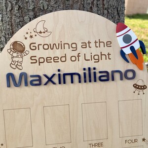 Pu&ograve; includere: Un grafico di crescita in legno a tema spaziale. Presenta un astronauta, un razzo, una luna crescente e stelle. Il testo recita "Growing at the Speed of Light Maximiliano" e ha quattro spazi vuoti per registrare le misure di altezza etichettati come "Three" e "Four".