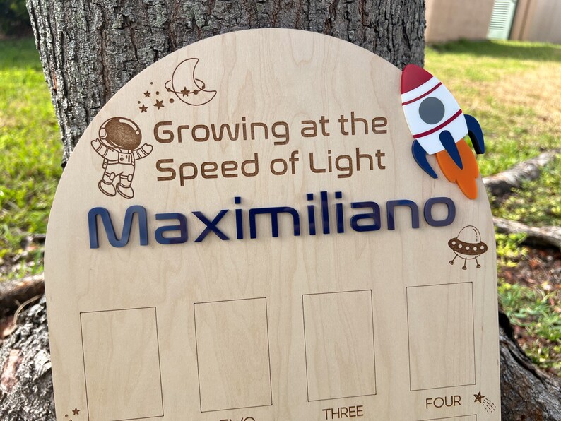 Pu&ograve; includere: Un grafico di crescita in legno a tema spaziale. Presenta un astronauta, un razzo, una luna crescente e stelle. Il testo recita "Growing at the Speed of Light Maximiliano" e ha quattro spazi vuoti per registrare le misure di altezza etichettati come "Three" e "Four".