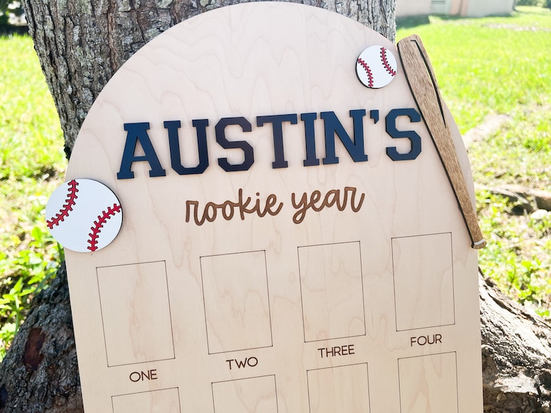 May include: A light-colored wooden arch-shaped sign featuring the text AUSTIN'S rookie year in dark blue and brown lettering. The sign is decorated with two white baseballs with red stitching and a wooden baseball bat, plus eight rectangular slots.