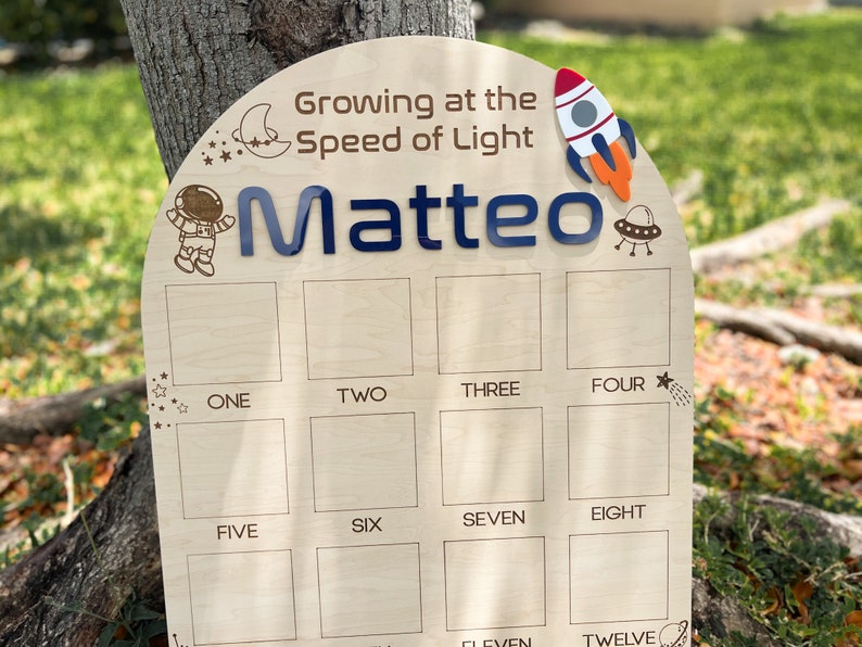 Pu&ograve; includere: Un grafico di crescita in legno a tema spaziale. Presenta un razzo, un astronauta, stelle e il testo "Growing at the Speed of Light Matteo". Il grafico ha 12 caselle vuote per registrare le misure di altezza.