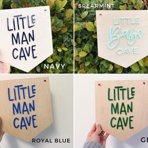 May include: Wooden pennant-shaped sign with the words "LITTLE MAN CAVE" in navy blue, royal blue, and green, and "LITTLE Babe CAVE" in spearmint. The signs are displayed against a background of greenery and a white wall.