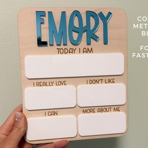 May include: A rectangular wooden milestone board featuring the name EMORY in metallic blue acrylic letters. The board includes the text TODAY I AM, I REALLY LOVE, I DON'T LIKE, I CAN, and MORE ABOUT ME, with blank white acrylic spaces for writing.