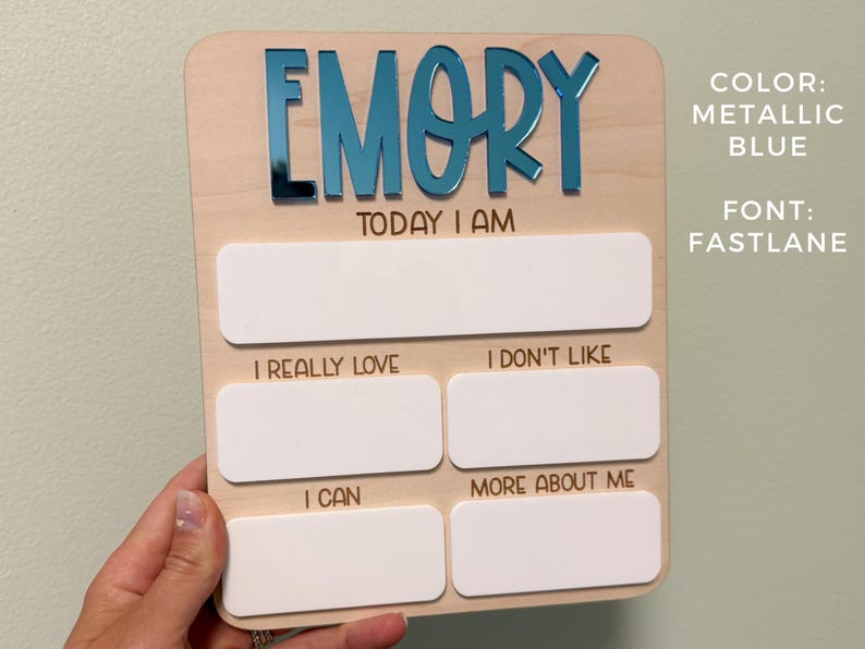 May include: A rectangular wooden milestone board featuring the name EMORY in metallic blue acrylic letters. The board includes the text TODAY I AM, I REALLY LOVE, I DON'T LIKE, I CAN, and MORE ABOUT ME, with blank white acrylic spaces for writing.