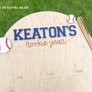 May include: A light-colored wooden plaque featuring the text "KEATON'S rookie year" in blue and brown, decorated with two white baseballs with red stitching and a wooden baseball bat cutout, including etched rectangular photo frames.