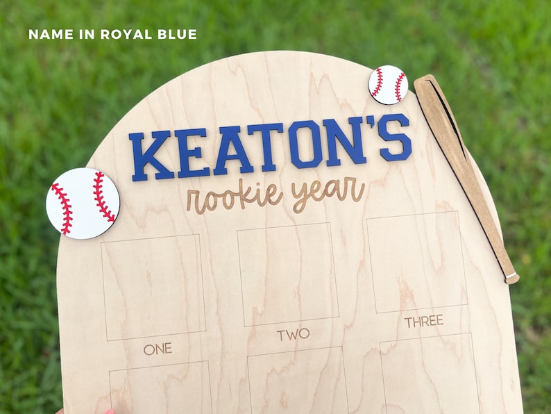May include: A light-colored wooden plaque featuring the text "KEATON'S rookie year" in blue and brown, decorated with two white baseballs with red stitching and a wooden baseball bat cutout, including etched rectangular photo frames.