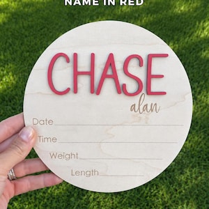 May include: A circular light-colored wooden birth announcement sign featuring the name CHASE in raised red letters and the name alan in script. Below the names are engraved lines with the labels Date, Time, Weight, and Length for recording details.