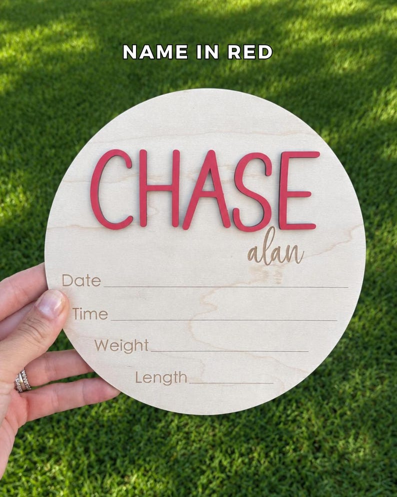 May include: A circular light-colored wooden birth announcement sign featuring the name CHASE in raised red letters and the name alan in script. Below the names are engraved lines with the labels Date, Time, Weight, and Length for recording details.