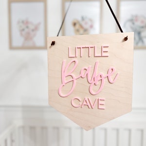 May include: A wooden pennant sign with the words "LITTLE Babe CAVE" in pink lettering. The sign is hanging from a black cord and is displayed in a nursery setting. Soft, neutral tones are used throughout the image.
