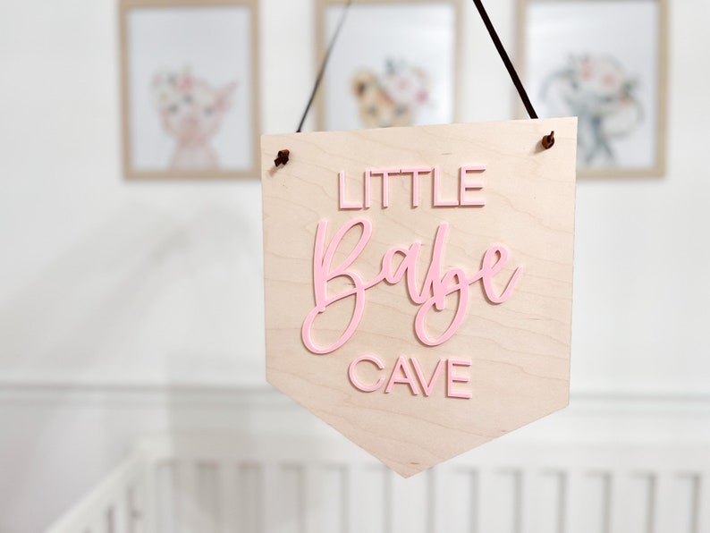 May include: A wooden pennant sign with the words "LITTLE Babe CAVE" in pink lettering. The sign is hanging from a black cord and is displayed in a nursery setting. Soft, neutral tones are used throughout the image.