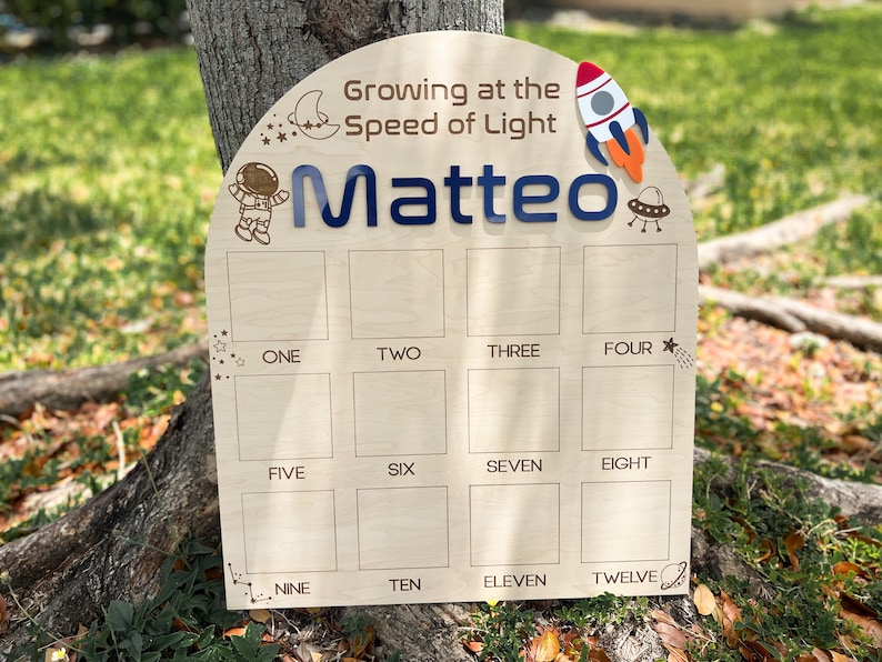 Pu&ograve; includere: Un grafico di crescita in legno a tema spaziale. Presenta un razzo, un astronauta, stelle e una luna crescente. Il grafico ha 12 spazi numerati per registrare l'altezza di un bambino. Il testo "Growing at the Speed of Light Matteo" &egrave; in cima al grafico.