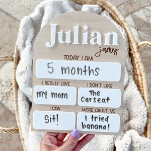 以下が含まれることがあります： 「Julian james」と名前が書かれた木製のマイルストーンボード。ボードは白地に白文字で飾られています。ボードは、年齢、好きなもの、新しいスキルなど、赤ちゃんのマイルストーンを追跡するために使用されます。ボードは、ジュリアンが5か月で、お母さんが大好きで、座ることができ、バナナを試したことを示しています。