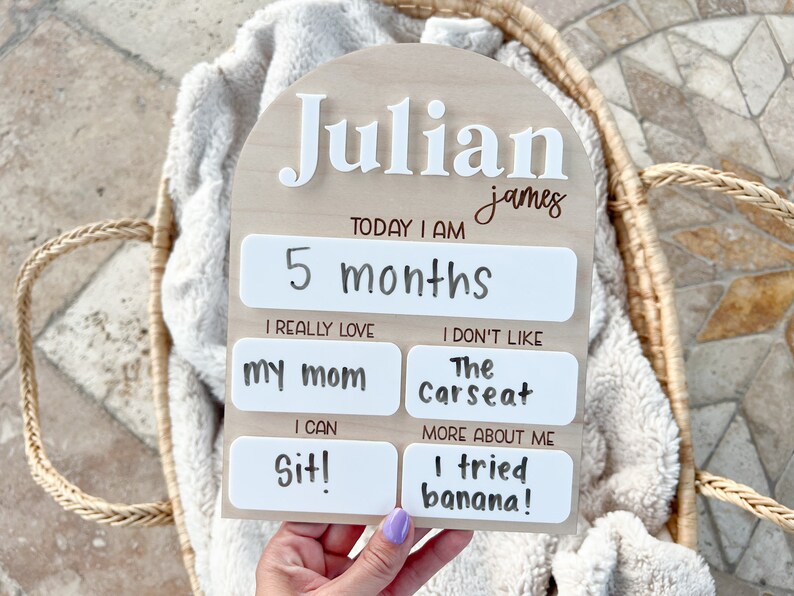 以下が含まれることがあります： 「Julian james」と名前が書かれた木製のマイルストーンボード。ボードは白地に白文字で飾られています。ボードは、年齢、好きなもの、新しいスキルなど、赤ちゃんのマイルストーンを追跡するために使用されます。ボードは、ジュリアンが5か月で、お母さんが大好きで、座ることができ、バナナを試したことを示しています。