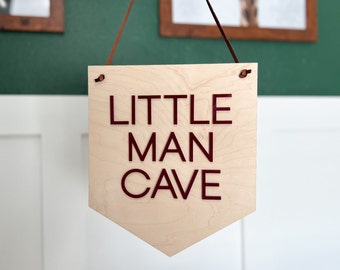 May include: A wooden sign with the words "Little Man Cave" in maroon lettering. The sign is hanging from a brown leather cord.
