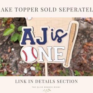 May include: A wooden cake topper featuring the text AJ IS ONE in navy blue and white layered lettering. The design includes a white baseball with red stitching and a wooden baseball bat graphic. The topper is attached to a thin wooden stick.