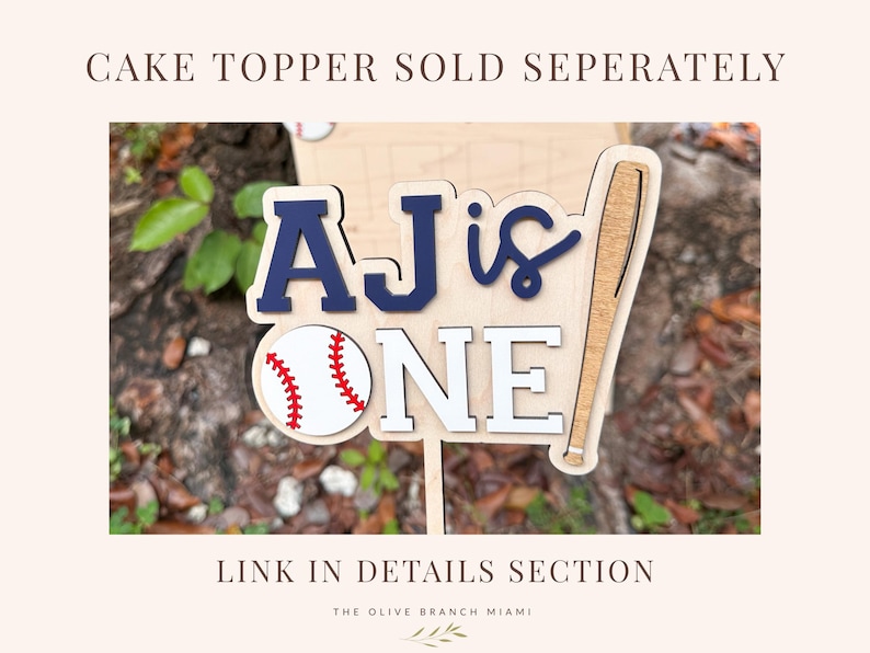 May include: A wooden cake topper featuring the text AJ IS ONE in navy blue and white layered lettering. The design includes a white baseball with red stitching and a wooden baseball bat graphic. The topper is attached to a thin wooden stick.
